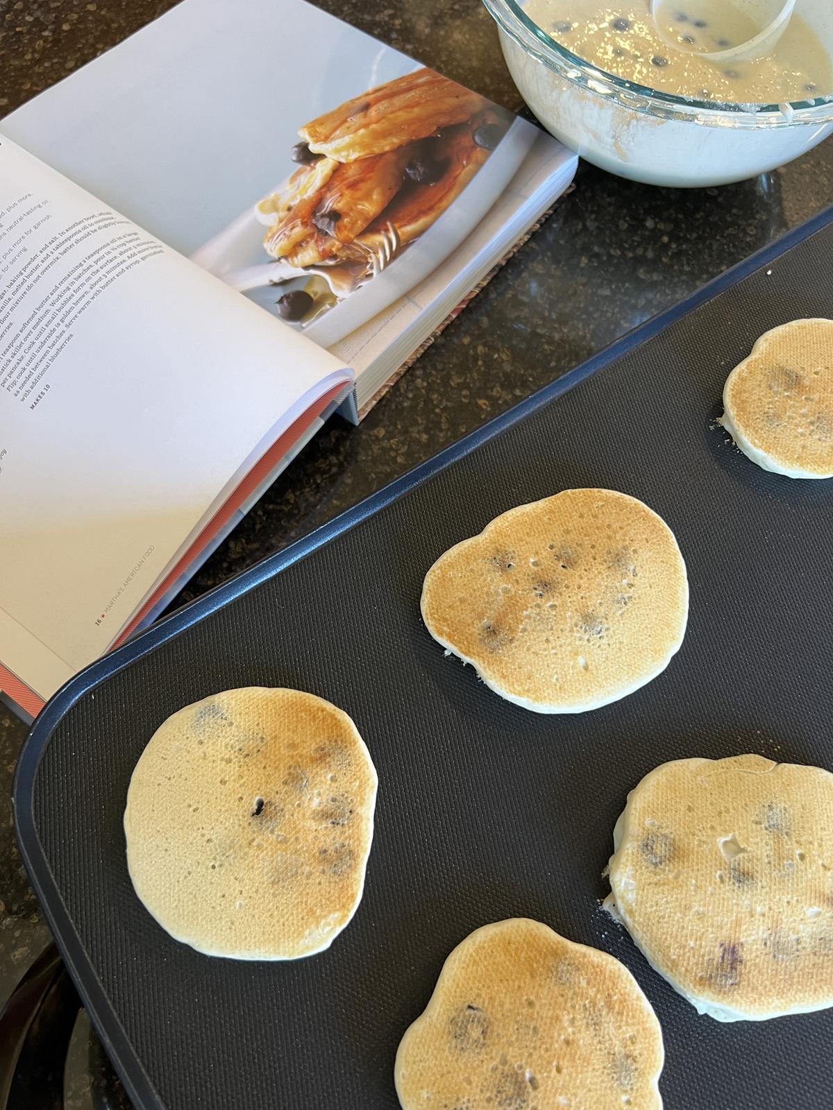 Blueberry pancakes cooking on griddle with cookbook and bowl of pancake batter in background