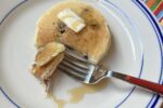 blueberry pancakes on white and blue plate with red fork and butter and syrup on pancake