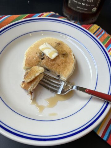 blueberry pancakes on white and blue plate with red fork and butter and syrup on pancake