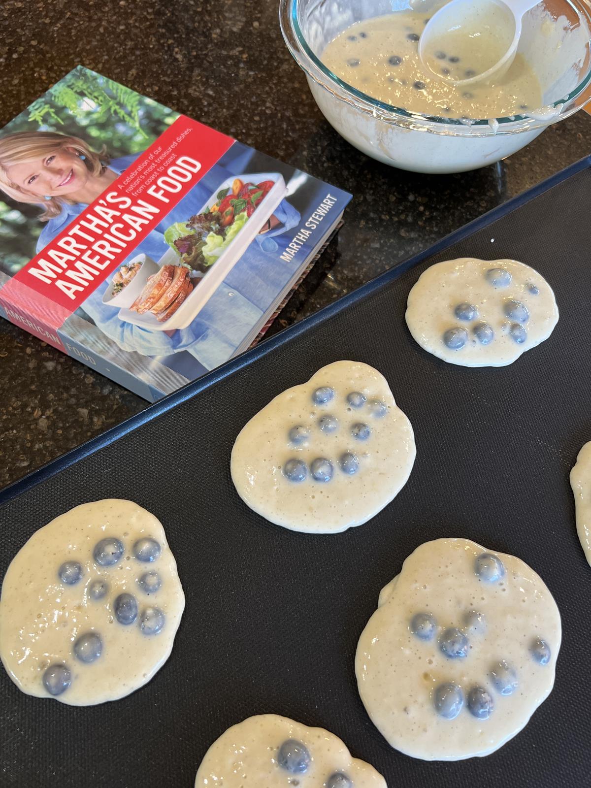 blueberry pancakes on griddle cooking with cookbook and bowl of pancake batter in background