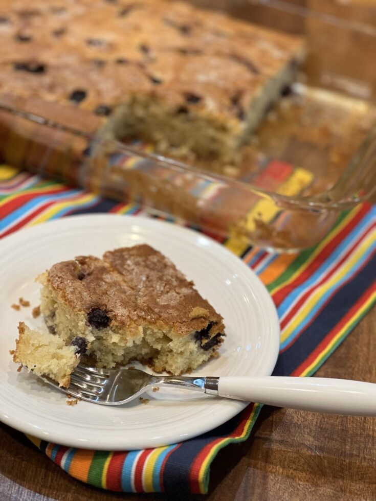 piece of blueberry coffee cake on white plate with white fork on striped napkin