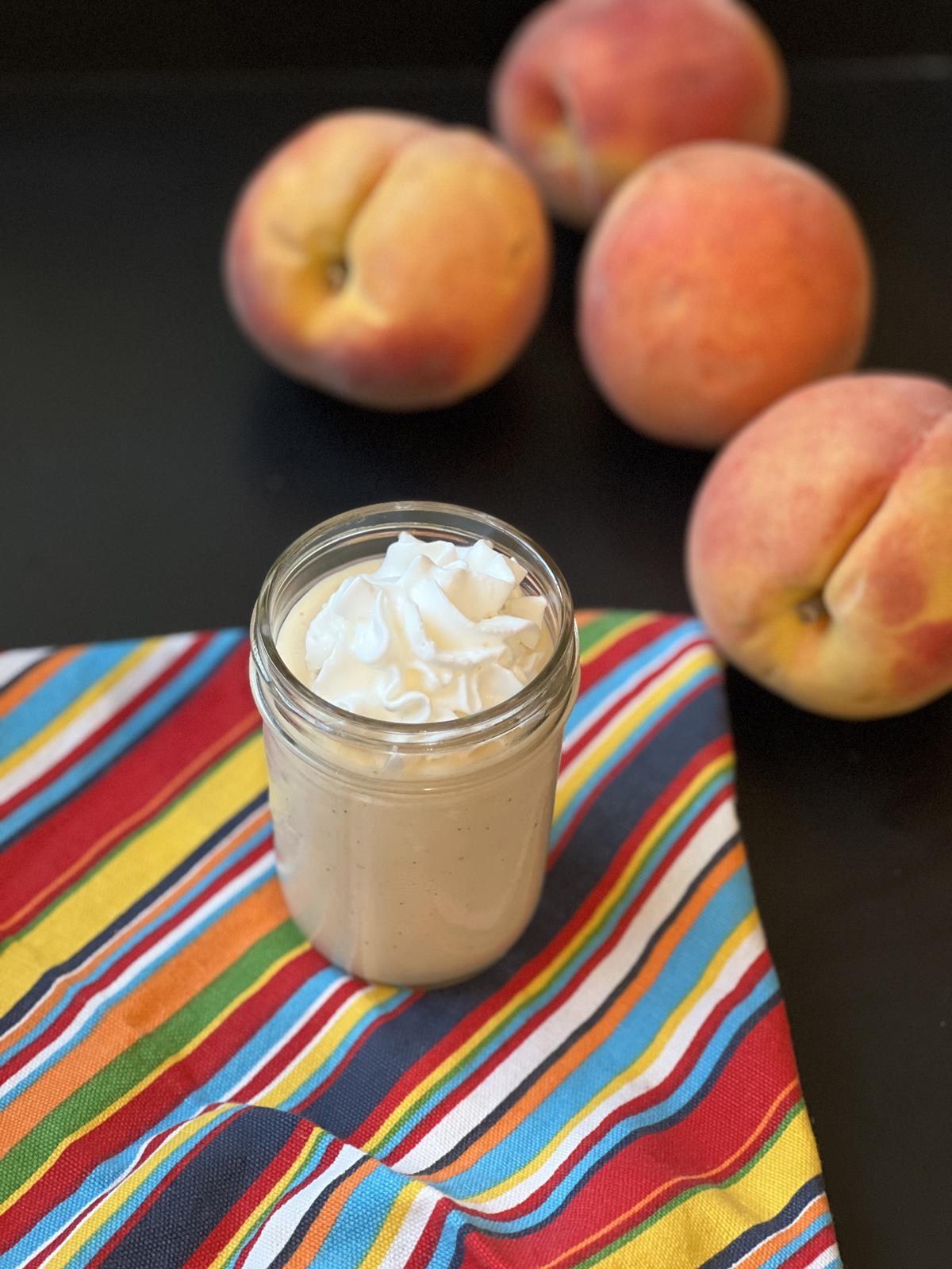 peach milkshake with whipping cream in glass jar cup with peaches in the background