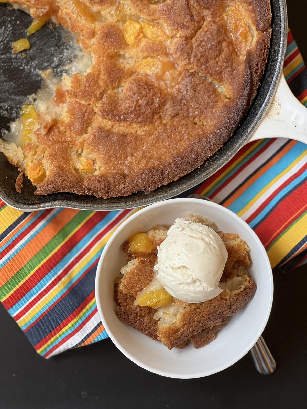 peach buckle in white bowl on striped napkin with pan of cobbler in background