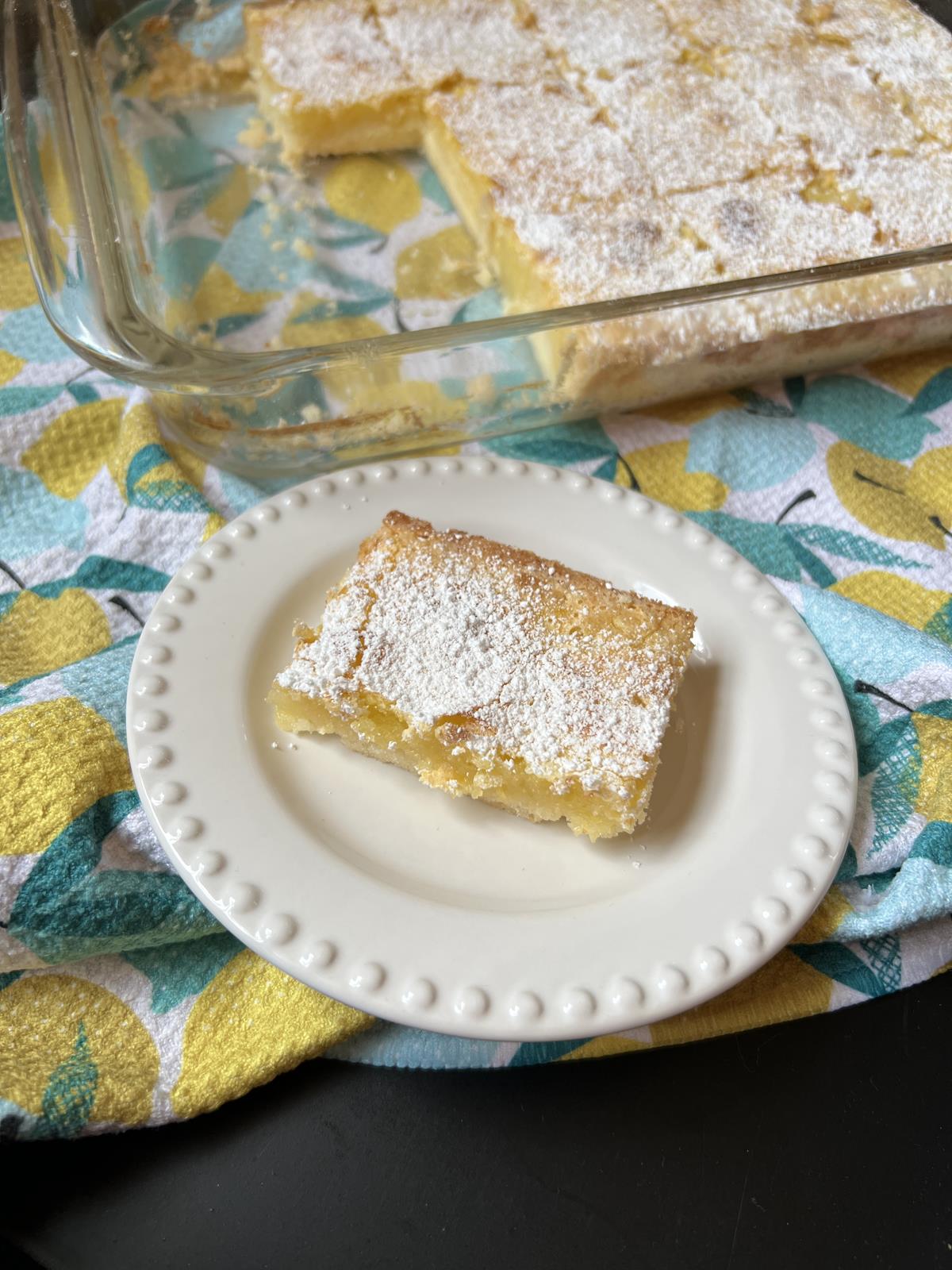 lemon bar on white plate with pan of lemon bars in background on napkin with lemons on it