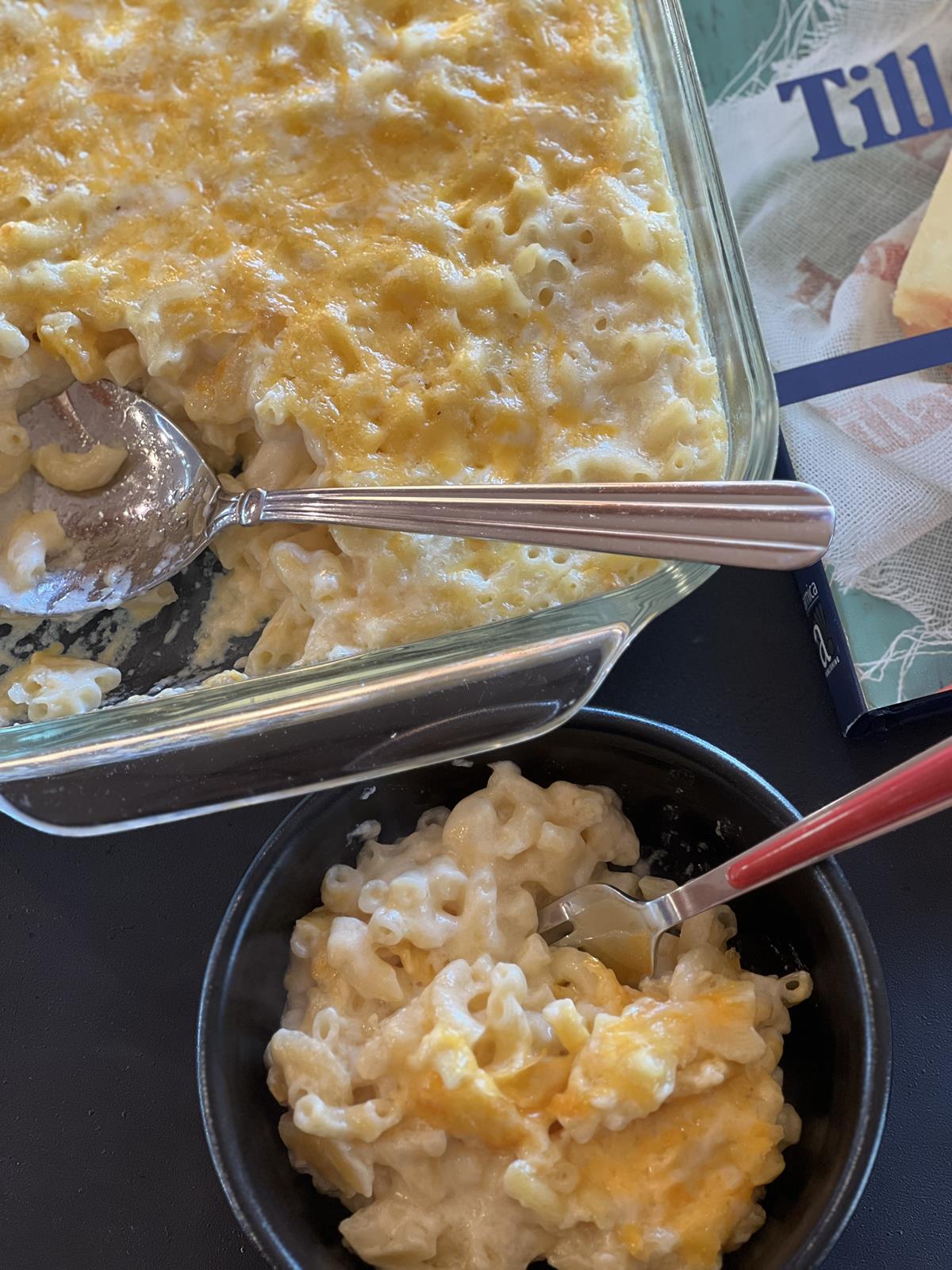 mac and cheese in black bowl with spoon and glass pan of mac and cheese in background