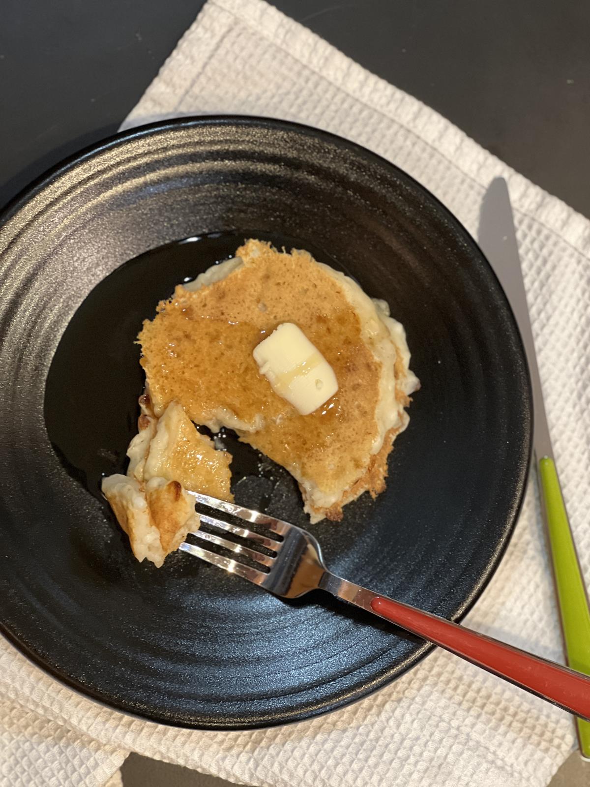 pancakes with butter and syrup on black plate with fork with red handle and knife with green handle