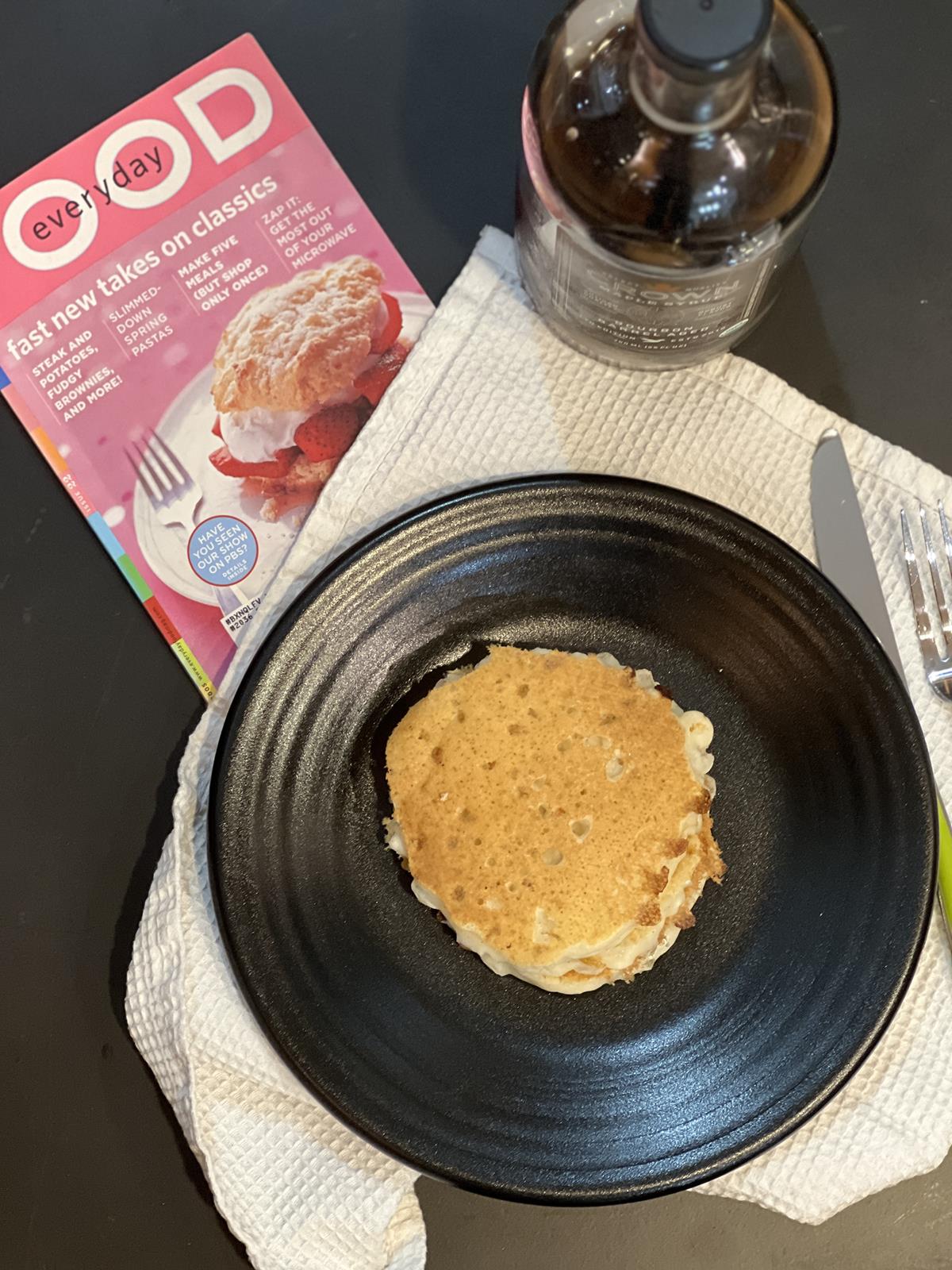 pancakes on black plate with knife and fork on white napkign with jar of maple syrup and a magazine in backgroumd