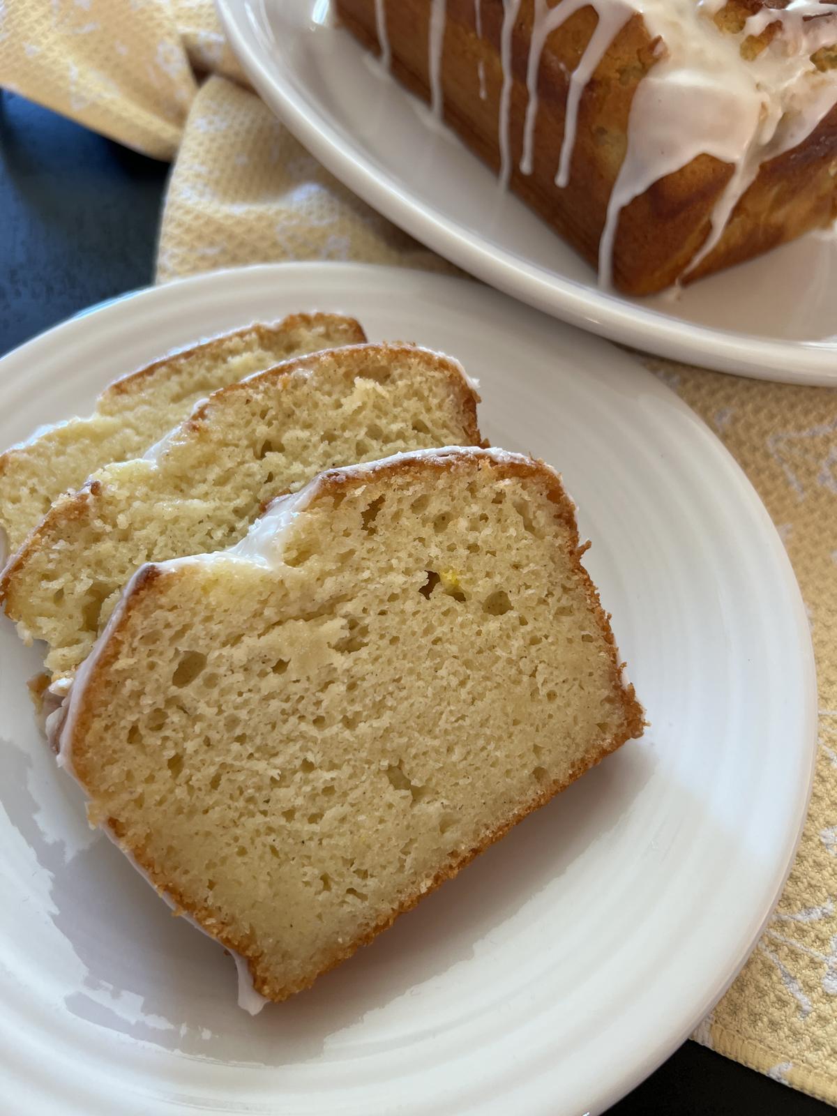 piece of lemon cake on white plate with loaf cake in background