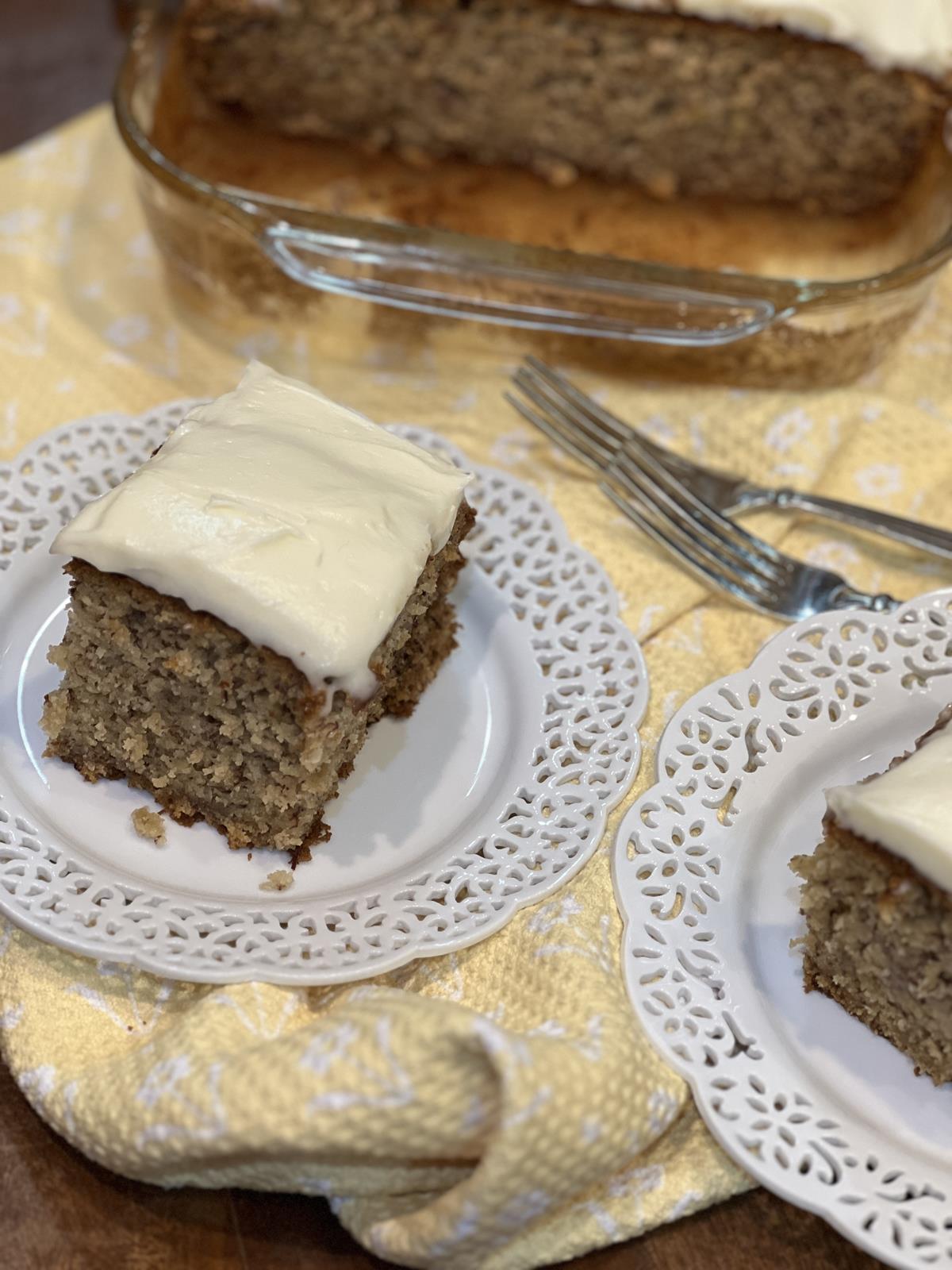 banana cake with frosting on two white plates with cake in glass pan in background