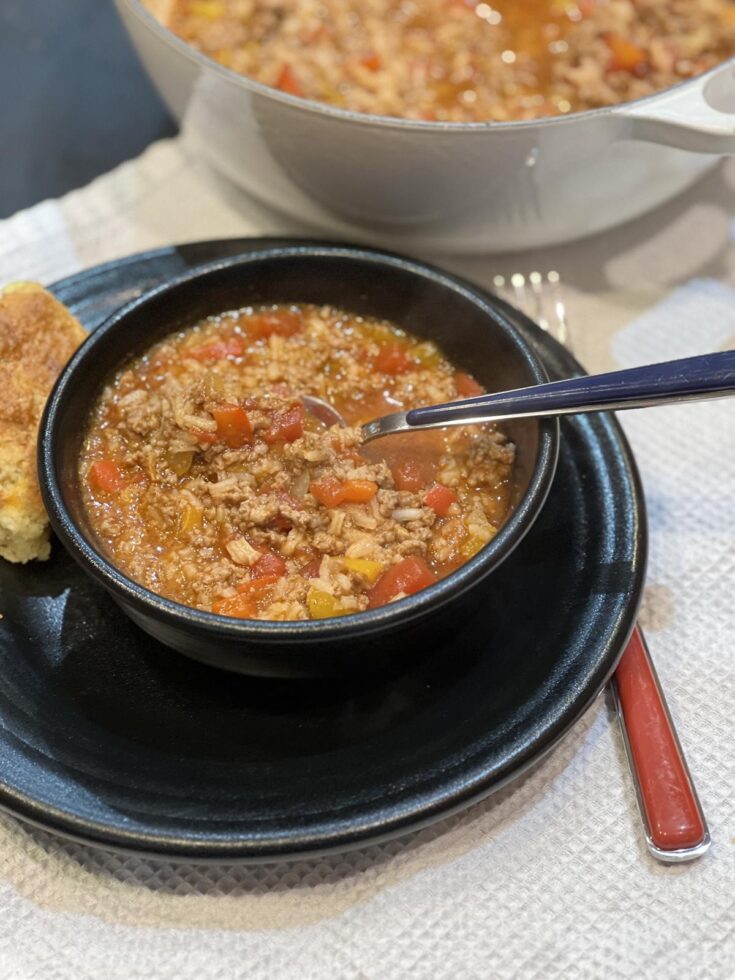 stuffed pepper soup in black bowl on black plate with biscuit and pan of soup in background