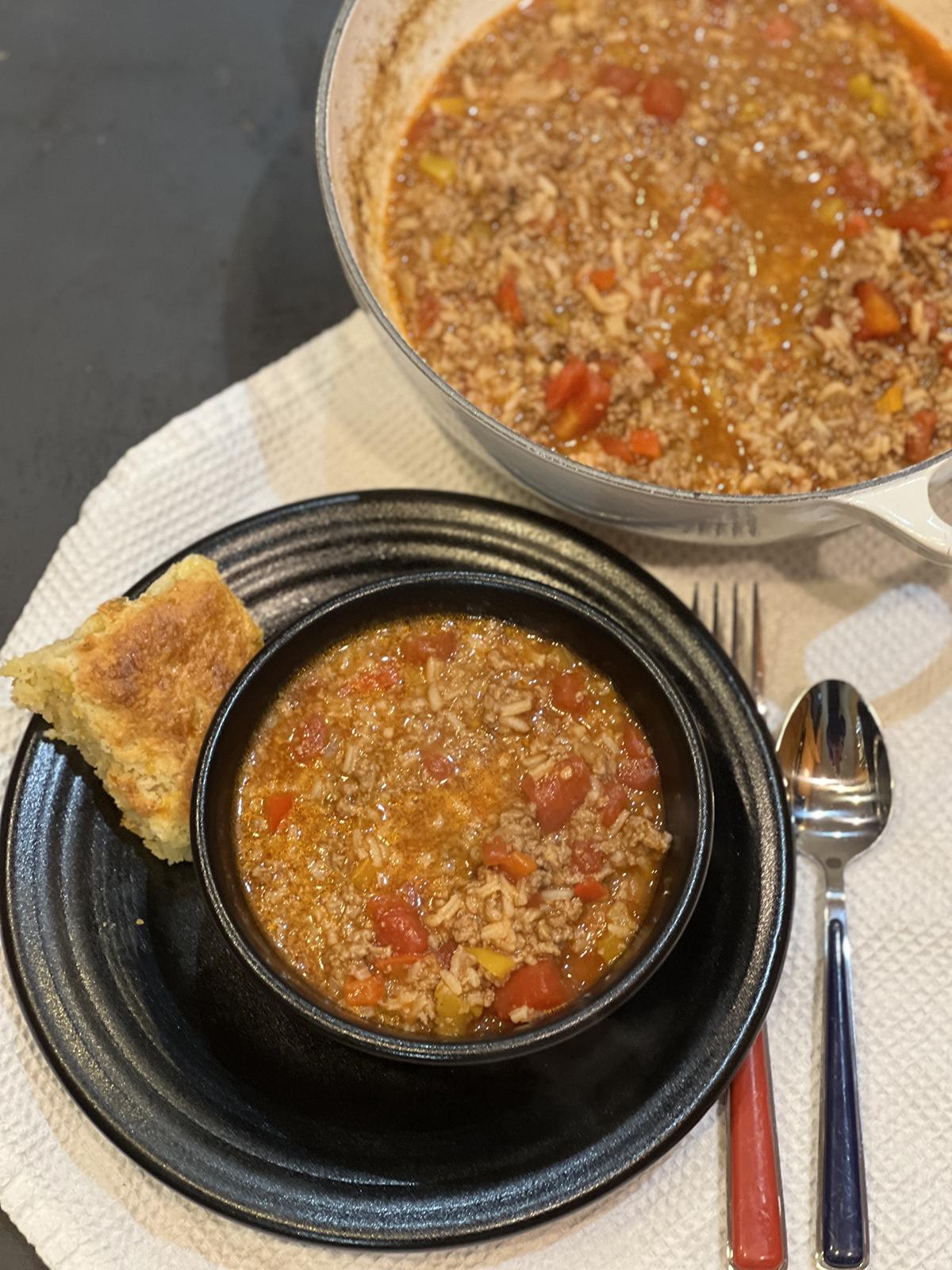 stuffed pepper soup in black bowl on black plate with biscuit and fork and spoon and white pot of soup in background