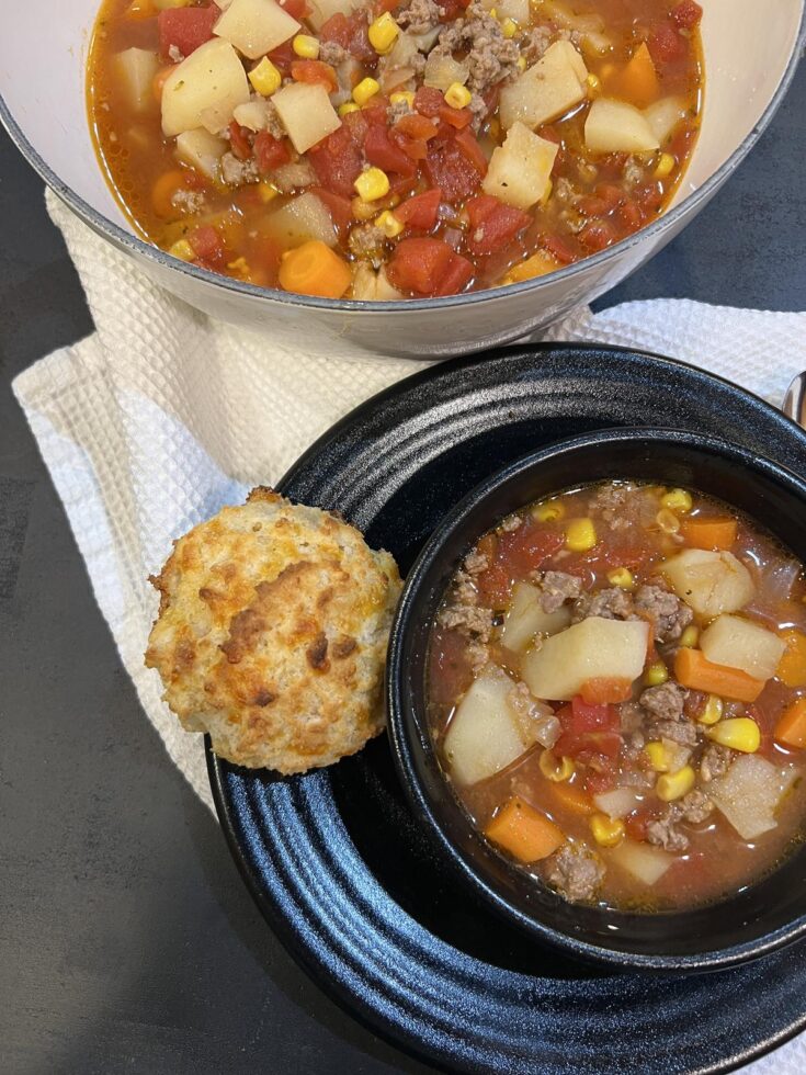 ground beef stew in white pot with bowl of stew in black bowl and biscuit on black plate