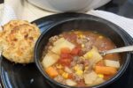 ground beef stew in black bowl with spoon and biscuit with pot of stew in background