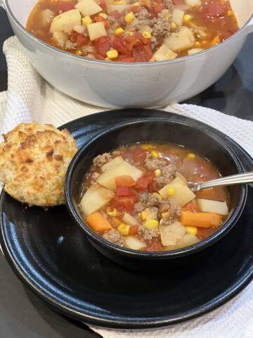 ground beef stew in black bowl with spoon and biscuit with pot of stew in background