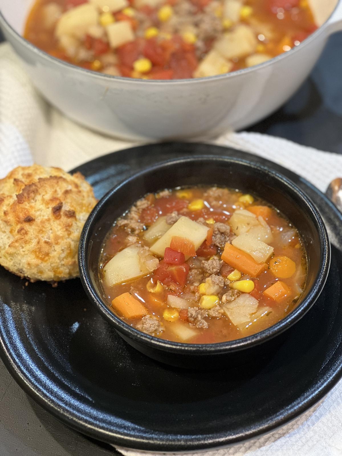 ground beef stew in black bowl on black plate with biscuit on the side