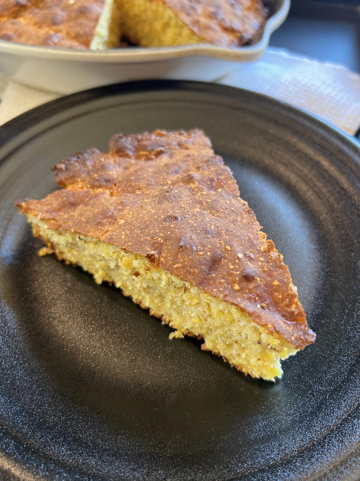 piece of Martha Stewart Cheddar Cornbread on black plate with pan of cornbread in background